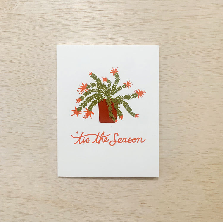Photo of holiday greeting card. Illustration of a Christmas cactus. Hand lettered text reads tis the season in red.